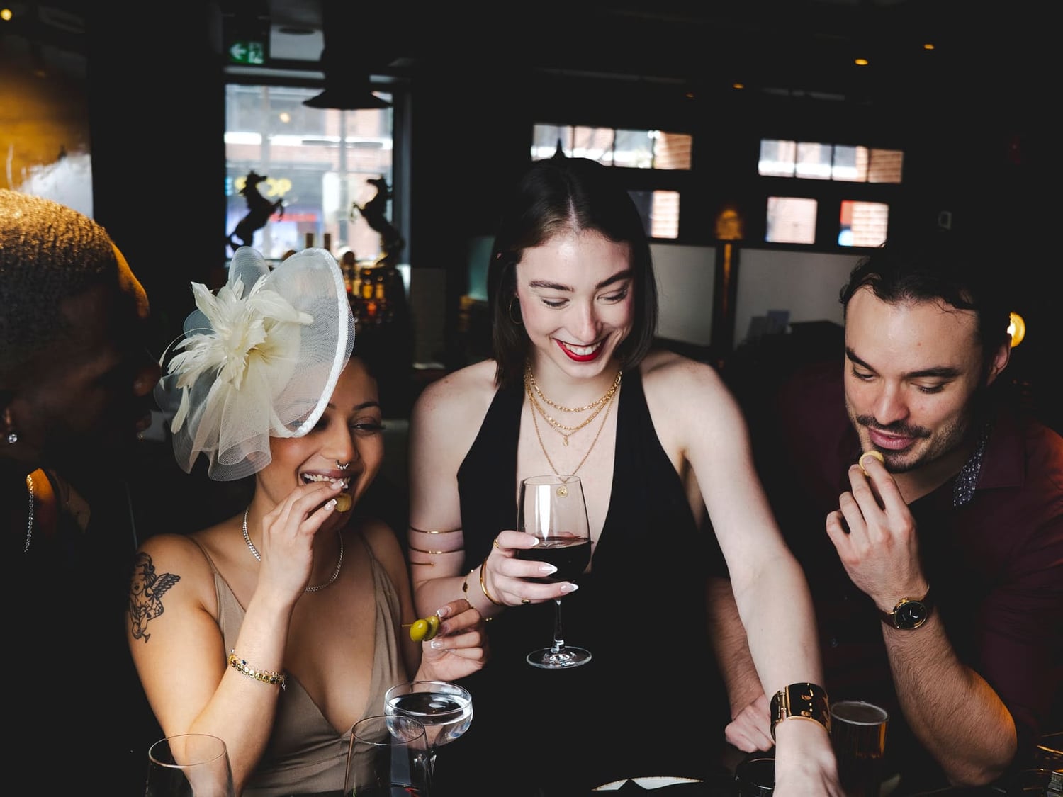 Four guests at a Notte Ristorante table mid-toast — a woman in a white fascinator laughing with a wine glass and olives in hand, the bronze lion silhouettes visible in the window behind