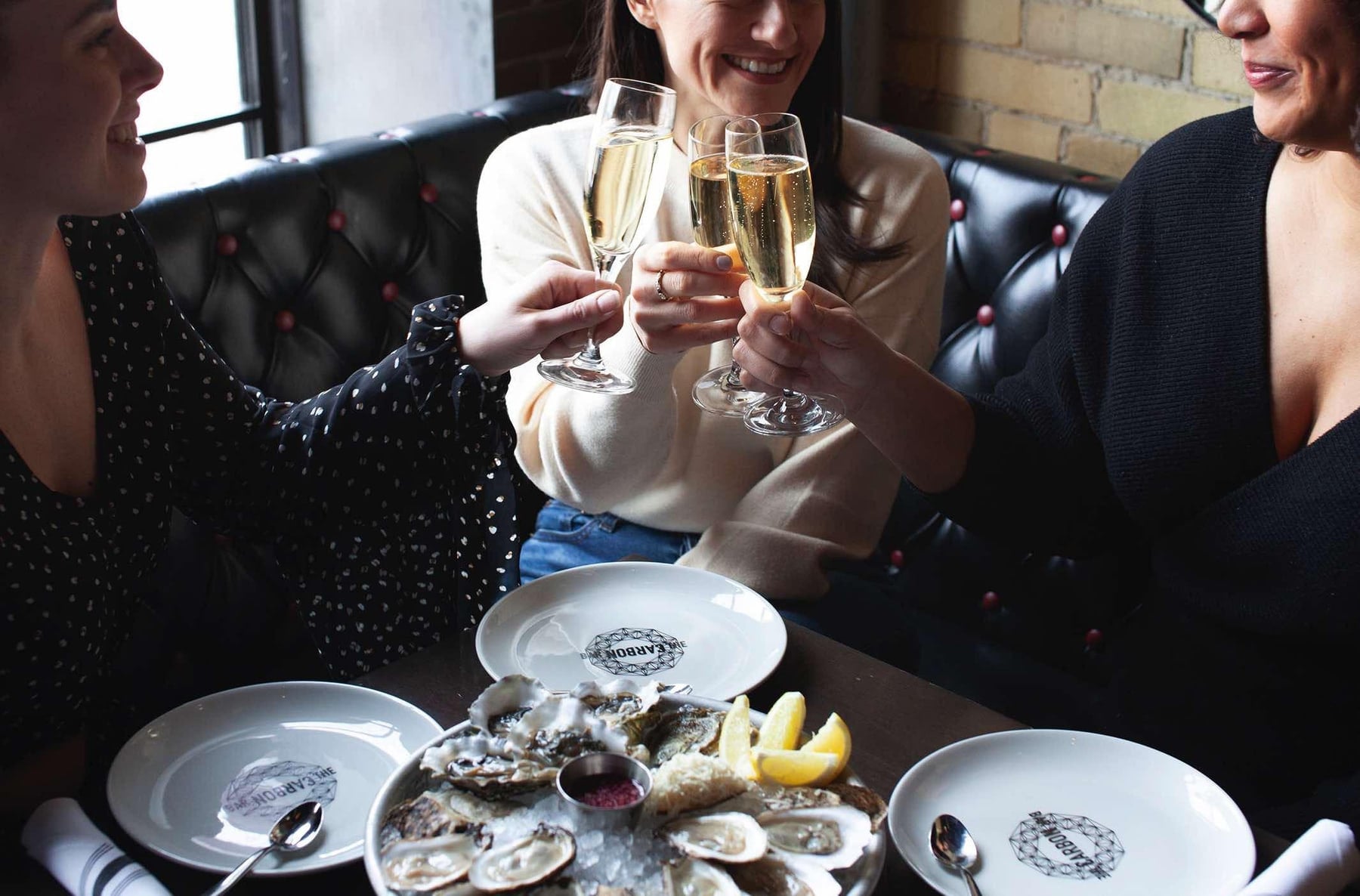 Three guests toasting with champagne flutes over a platter of oysters at a Carbon Bar booth — tufted black leather banquette, branded Carbon Bar plates, exposed brick wall behind