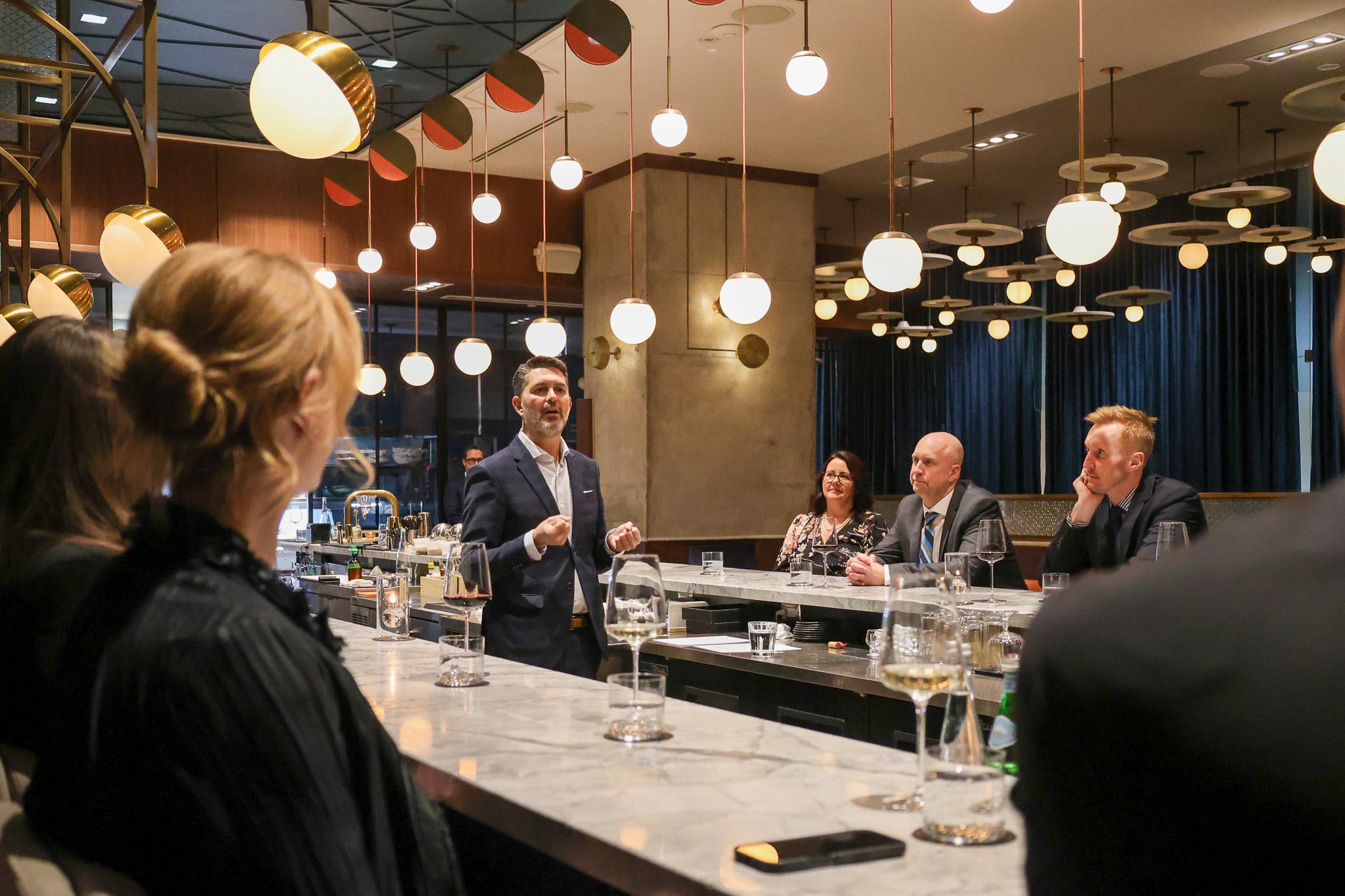 Yannick Bigourdan at table in a polished dining room, raising glass with guests — the choreography of hospitality in frame