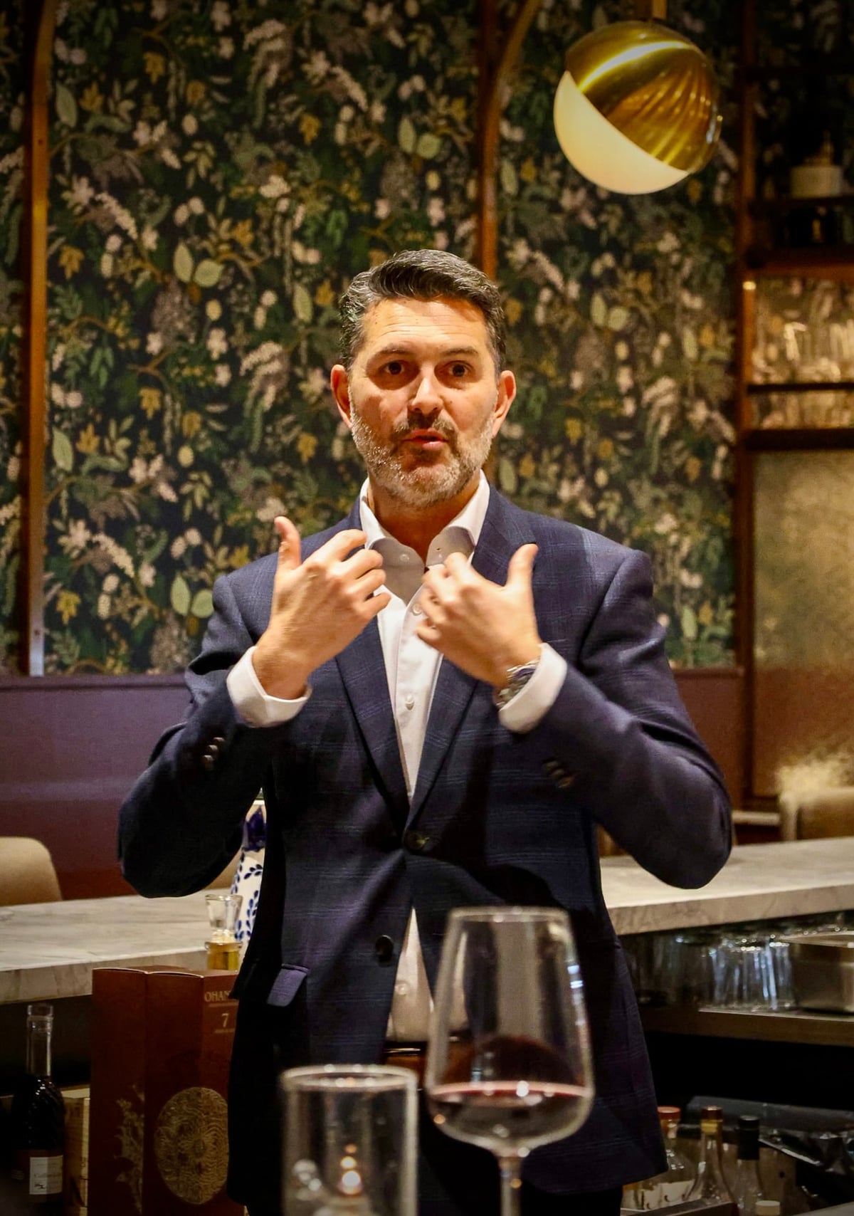 Yannick Bigourdan teaching at a marble bar — explaining a point to guests in a fine dining room with botanical-papered walls, a glass of red wine on the counter in foreground