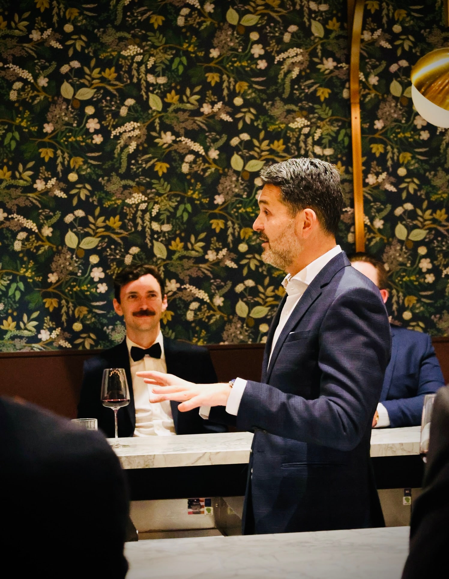 Yannick Bigourdan speaking at a private dining table with floral wallpaper, a bow-tied guest watching — the architecture of the room being taught, not described