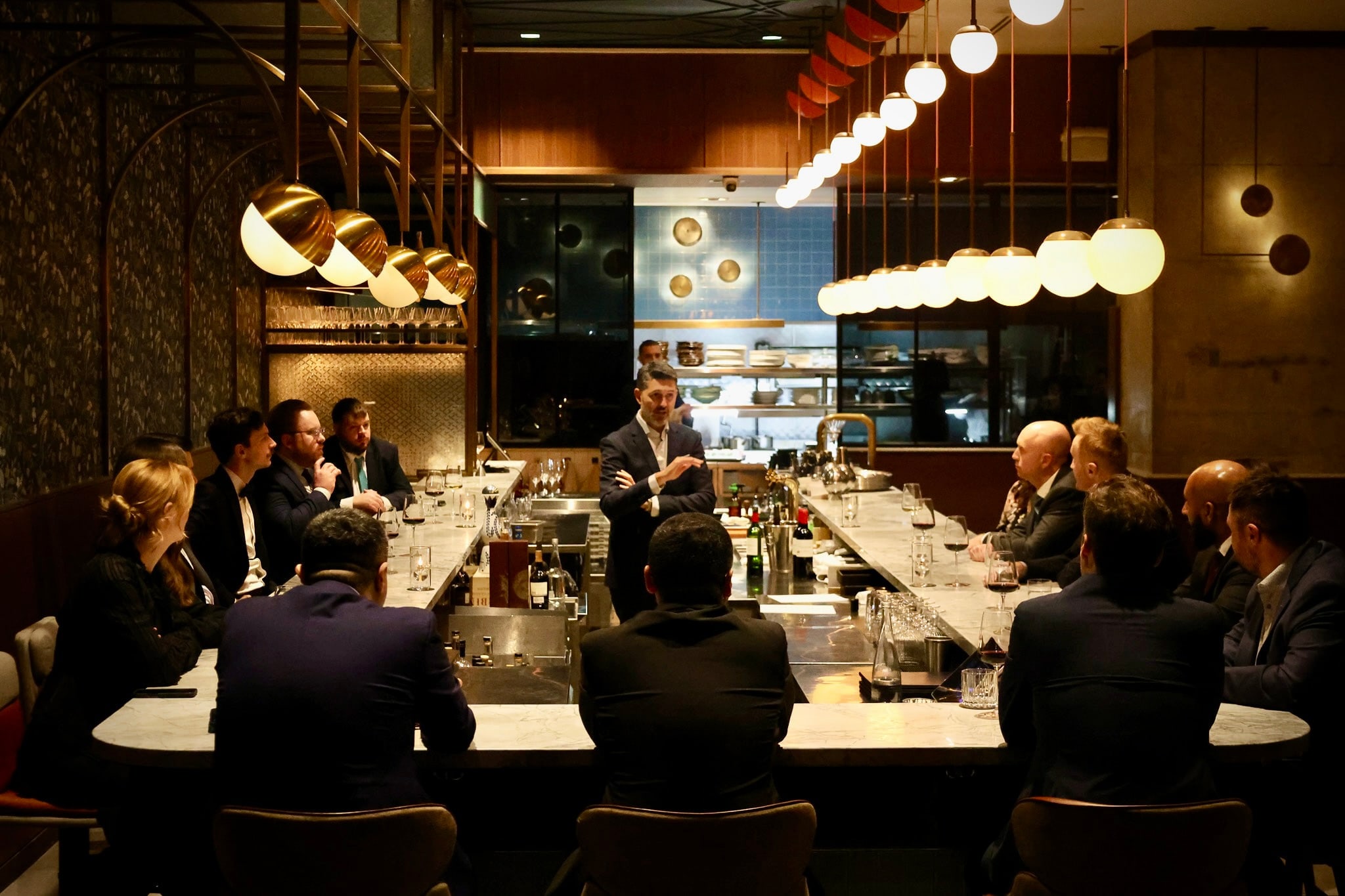 A working dinner at a marble bar — Yannick Bigourdan at the head, addressing a full table of operators and founders, pendant lights overhead