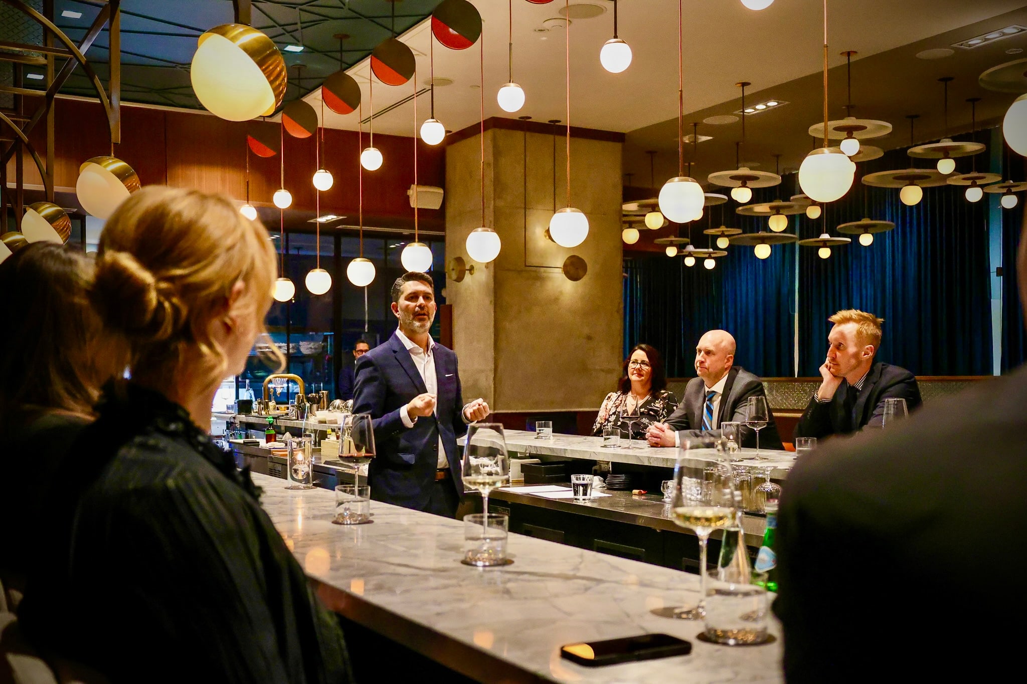 Yannick Bigourdan addressing a full room of operators and founders from the head of a marble bar — pendant lights overhead, a working dinner in evening attire