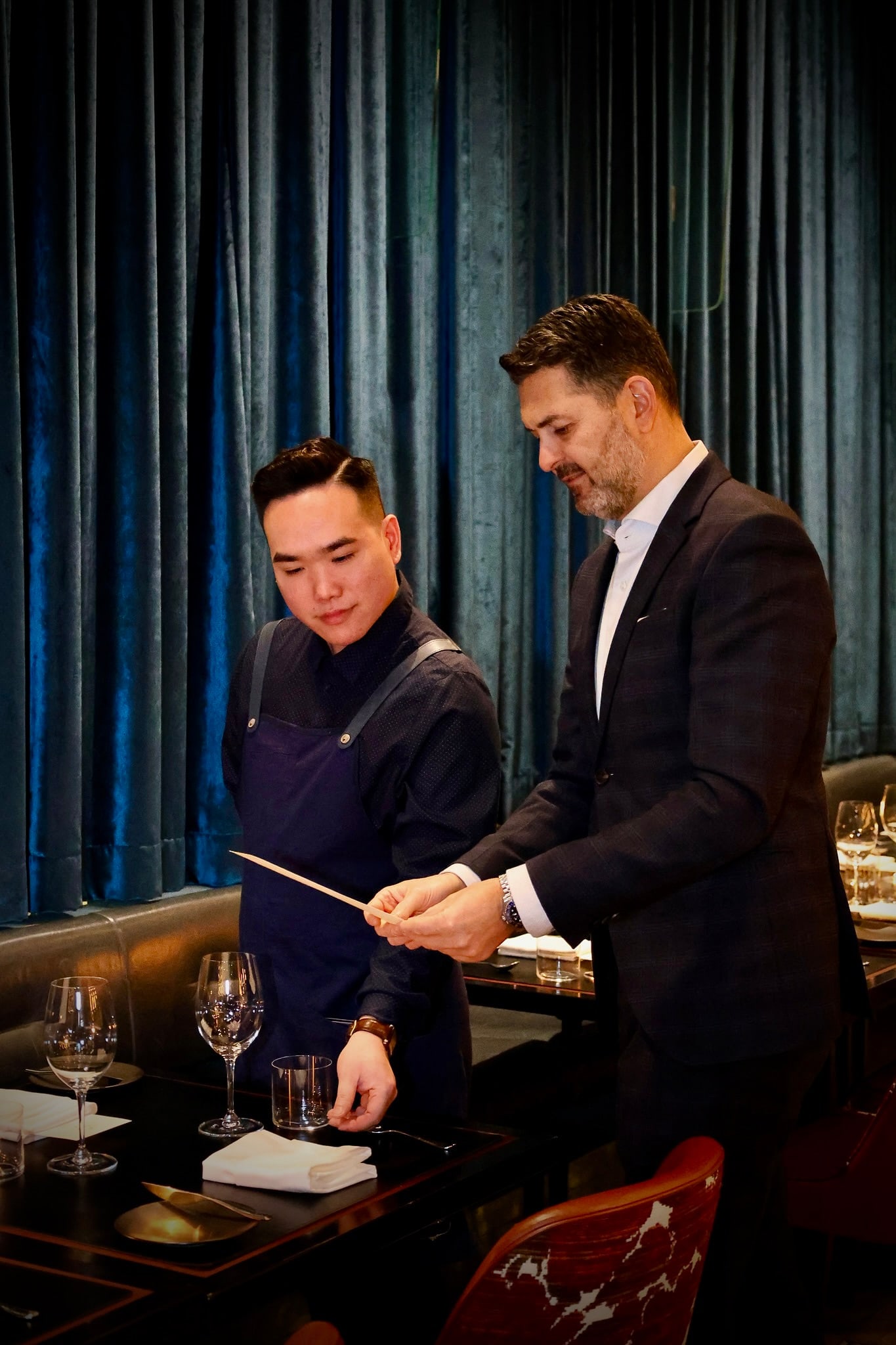 Yannick Bigourdan walking a young sommelier through a table-setup detail in a fine dining room, blue velvet curtains behind — a peer on the floor, mid-explanation